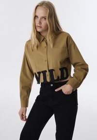 Camisa verde oliva cortada de botones con un bolsillo frontal y letras negras "WILD" en el dobladillo, combinada con jeans negros de cintura alta.