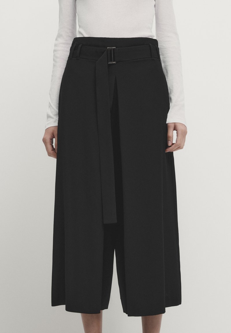 Femme portant un pantalon noir taille haute, jambes larges et coupé au-dessus de la cheville, avec une ceinture à l'avant et une boucle argentée, sur un haut blanc à manches longues.