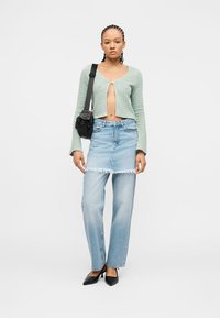 Νεαρή γυναίκα που φοράει πράσινη κοντή ζακέτα, στρωμένη ανοιχτόχρωμη τζιν φούστα πάνω από τζιν παντελόνι, μαύρα μυτερά τακούνια και μαύρη τσάντα ώμου, όρθια.