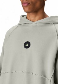 Lichtgrijze hoodie met een grote zwarte cirkelvormige patch met drie groene stippen. Het heeft een losse pasvorm en rafelige zoomdetails.