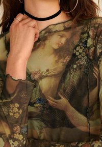 En gennemsigtig bluse med et blomster- og klassisk maleri print, med en scalloped halsudskæring og lange, flydende ærmer. Sort choker-accessory synlig.