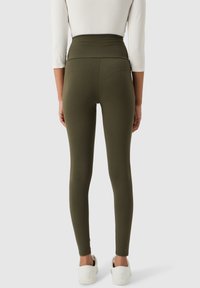 Leggings vert olive avec une taille haute, fabriqués à partir d'un tissu extensible, offrant une texture lisse et un design ajusté, associés à des chaussures blanches.