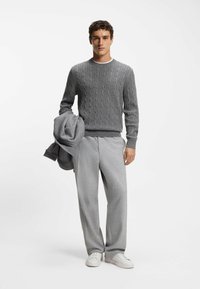 Grauer Kabelstrickpullover mit gerippten Bündchen und Saum, kombiniert mit hellgrauen, maßgeschneiderten Hosen und weißen Sneakers, hält eine graue Jacke.