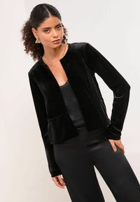 Černá sametová zkrácená bunda s dlouhými rukávy, otevřeným předním dílem a dvěma předními kapsami. Spárována s černým saténovým topem a kalhotami.
