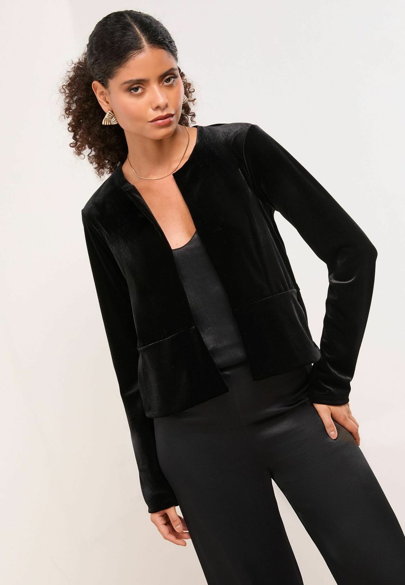 Černá sametová zkrácená bunda s dlouhými rukávy, otevřeným předním dílem a dvěma předními kapsami. Spárována s černým saténovým topem a kalhotami.