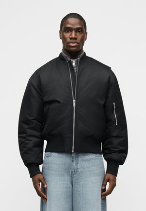 Mann trägt eine schwarze Bomberjacke mit Reißverschluss-Detail und hellblaue Jeans, steht vor einem schlichten, hellen Hintergrund.