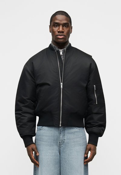Mann trägt eine schwarze Bomberjacke mit Reißverschluss-Detail und hellblaue Jeans, steht vor einem schlichten, hellen Hintergrund.