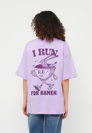 Persona con capelli ricci che indossa una maglietta lavanda che mostra un cartone animato di una ciotola di ramen che cammina con le bacchette e il testo "Corro per il ramen."