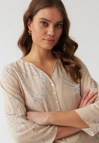 Beige Bluse mit langen Ärmeln, die ein dezentes florales Muster in Hellgrau aufweist, sowie einen V-Ausschnitt und Frontknöpfe. Weiche, glatte Textur.