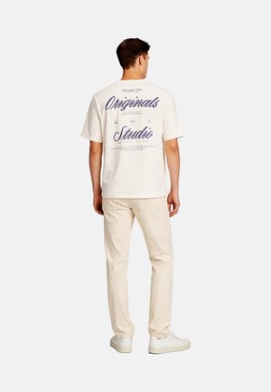 Uomo in piedi di spalle, con una t-shirt bianca con scritta blu, pantaloni beige e sneakers bianche su sfondo semplice.