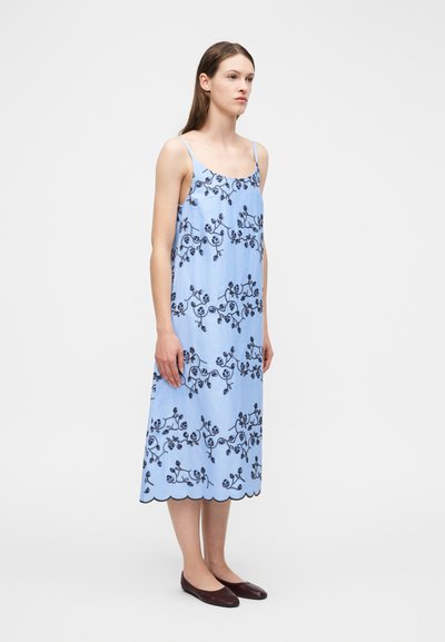 Γυναίκα που φοράει αμάνικο γαλάζιο midi φόρεμα με σκοτεινή ανθισμένη κεντητική και κυματιστό τελείωμα, συνδυασμένο με σκοτεινά επίπεδα παπούτσια, στέκεται πλαγίως.