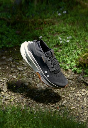 Zwarte hardloopschoen met witte zool, gelabeld "Nike ZoomX", boven rotsachtige en grasrijke grond, zwevend in de lucht met modderdeeltjes eronder.