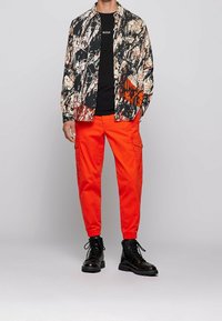 Blusenhemd mit floralem Muster in Schwarz und Beige über einem schwarzen T-Shirt; kombiniert mit leuchtend orangefarbenen Cargo-Hosen und schwarzen Schnürstiefeln.