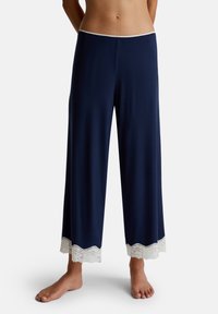 Marineblauwe wijde pyjamabroek met witte kantafwerking aan de enkels, gedragen op blote voeten door een staande persoon tegen een eenvoudige witte achtergrond.