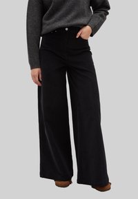 Zwarte wijde pijpen broek gemaakt van zachte stof, met een hoge taille, voorzakken en minimale hardware. Gecombineerd met een grijze trui.