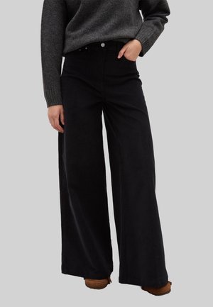 Pantaloni neri a gamba larga in tessuto morbido, con vita alta, tasche frontali e dettagli hardware ridotti al minimo. Abbinati a un maglione grigio.