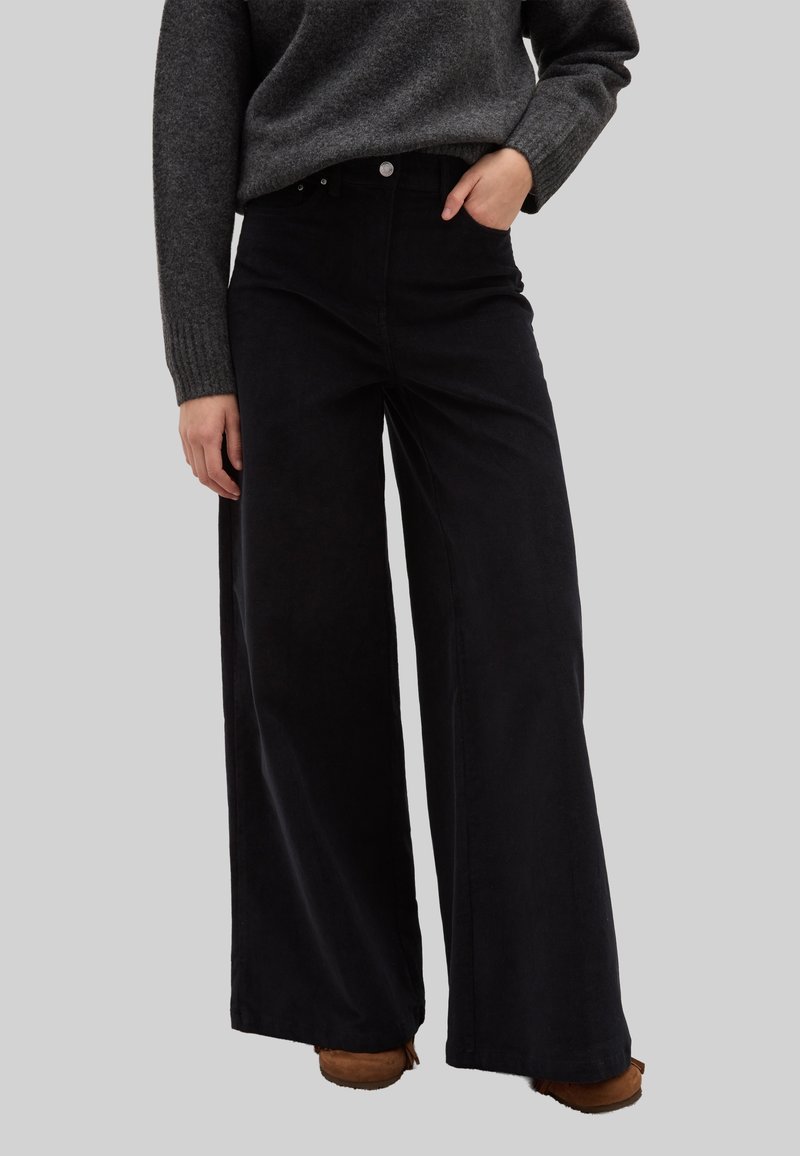 Zwarte wijde pijpen broek gemaakt van zachte stof, met een hoge taille, voorzakken en minimale hardware. Gecombineerd met een grijze trui.