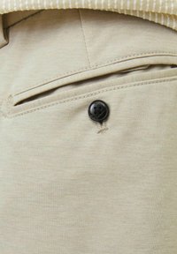 Beige színű pamut nadrág, hátoldalán egy díszes fekete gombbal ellátott zsebbel. Az anyag sima textúrájú, finom színváltozásokkal.