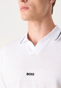 Biely polo tričko s mriežkovou štruktúrou, s čiernym pruhom pozdĺž goliera a výrazným čiernym logom "BOSS" na hrudi.
