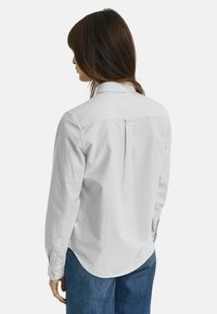 Donna con capelli castani che indossa una camicia a maniche lunghe grigia chiara a righe e jeans blu, vista di spalle su uno sfondo bianco.