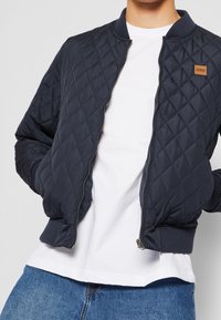 Námořnická prošívaná bomber bunda s ribovaným límcem a manžetami, vyznačující se zipovým zapínáním a malou koženou etiketou na hrudi.