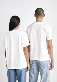 Bílé bavlněné trička, krátké rukávy, volný střih, pohled zezadu. Kombinace se světlými modrými džíny, minimalistický design, bez viditelných log. 