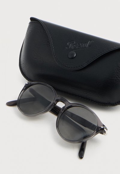 Μαύρα στρογγυλά γυαλιά ηλίου με σκούρους φακούς που ακουμπούν δίπλα σε μαύρη δερμάτινη θήκη Persol πάνω σε λευκή επιφάνεια.