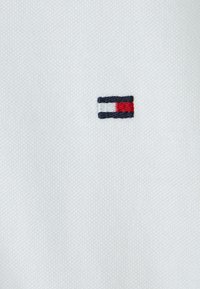 Biała tkanina o teksturowanej powierzchni z małym, haftowanym logo w kolorze granatowym, czerwonym i białym, umieszczonym po lewej stronie materiału.