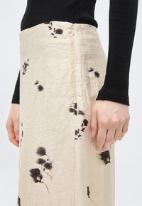 Μπεζ φούστα με μαύρα λουλουδάτα σχέδια, υφασμάτινη υφή, πλαϊνό φερμουάρ· άτομο που φοράει μαύρο τοπ με μακριά μανίκια, με το χέρι ακουμπισμένο στο πλάι.