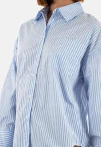 Camicia button-up a righe blu chiaro e bianche, con colletto, maniche lunghe e una tasca sul petto. Tessuto morbido con una vestibilità rilassata.