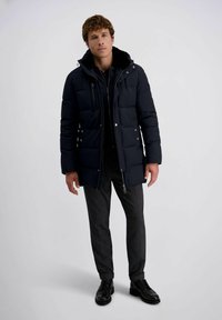 Man staat in een marineblauwe gewatteerde winterjas, donkergrijze broek en zwarte nette schoenen tegen een effen lichte achtergrond.