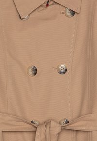 Beige trenchcoat med dobbeltbrystet design, store gjennomsiktige knapper og belte med knyting. Glatt tekstur og synlige sømdetaljer.