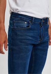 Blå denimjeans med rak skärning, femficksdesign, subtil blekning och kontrasterande sömnadsdetaljer. Metallknapp vid midjan.