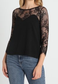 Top nero con maniche lunghe in pizzo trasparente con motivi floreali e corpino nero solido. Collo rotondo, vestibilità rilassata e texture morbida.