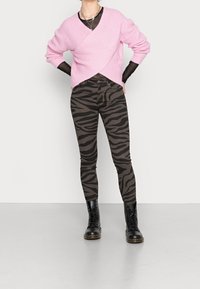 Różowy sweter prążkowany z dekoltem w serek, noszony na czarnej bluzce z długim rękawem. Brązowe dopasowane spodnie z czarnym nadrukiem zebry oraz czarne buty sznurowane.