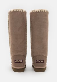 Bottines en suede marron montantes jusqu'au genou avec des coutures beiges et un accent texturé sur le côté, présentant une semelle marron et un patch logo en violet foncé.