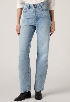 Personne portant un jean bleu clair taille haute à jambes droites, une chemise blanche à manches longues retroussées et des bottines noires, debout sur un fond uni.