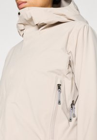 Jachetă impermeabilă bej deschis, cu glugă cu guler înalt, buzunare cu fermoar și șnururi gri, având o textură netedă, mată.