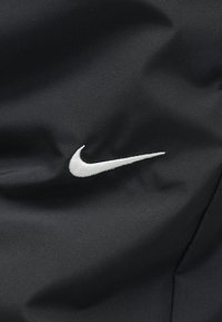 Μαύρο ύφασμα με ομαλή υφή που φιλοξενεί λευκό κεντητό λογότυπο Nike swoosh. Χωρίς πρόσθετα μοτίβα ή εξαρτήματα ορατά.