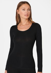 Vrouw in een aansluitend zwart shirt met lange mouwen en een ronde halslijn, die iets opzij kijkt tegen een effen witte achtergrond.