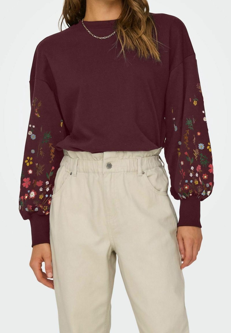 Femme portant un haut bordeaux à manches longues avec des motifs floraux sur les manches et un pantalon taille haute beige clair.