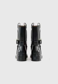 Botas de cuero negras hasta el tobillo con una textura suave, dos correas con hebillas y tiradores en la parte trasera. Tacones cuadrados y logo embossado en la suela.
