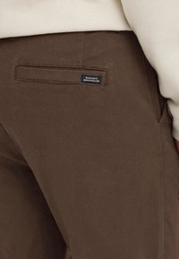 Brune bukselomme med liten svart etikett som leser "Dedikert til denimperfeksjon" på en person iført kremfarget topp.