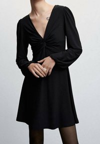 Vestido negro con mangas largas, escote en V y detalle frontal retorcido. Falda fluida y textura suave. Usado sobre medias negras.