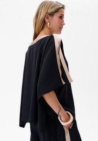 Zwarte tuniek met brede mouwen, voorzien van beige zijstrepen. Lichtgewicht stof, losse pasvorm en een geaccentueerde arm met een decoratieve armband.