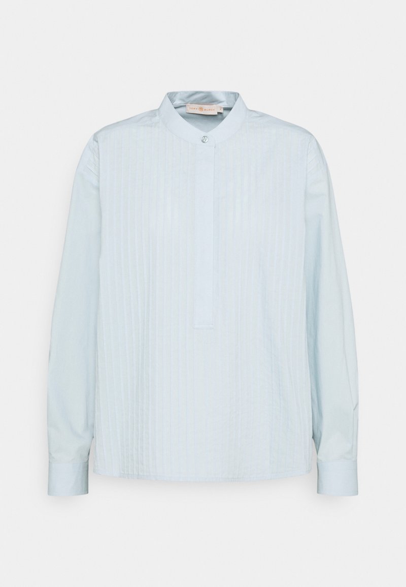 Blouse bleu clair à manches longues avec plis verticaux à l’avant, col officier avec un bouton unique et patte de boutonnage cachée.