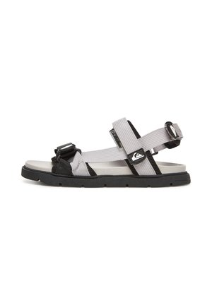 Grå og sort sandal med åben tå med justerbare stofremme og sort gummisål, vist fra siden på en hvid baggrund.