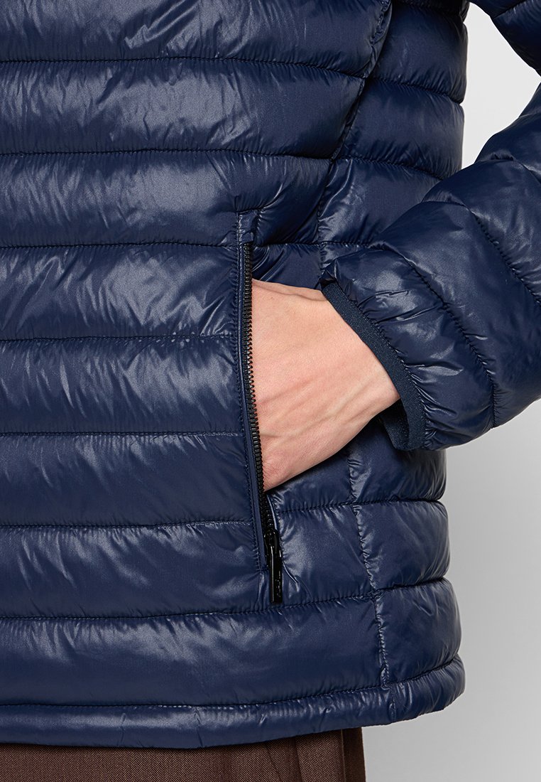 Mano introducida en el bolsillo lateral con cremallera de una chaqueta acolchada azul marino, con costuras horizontales y puños elásticos.