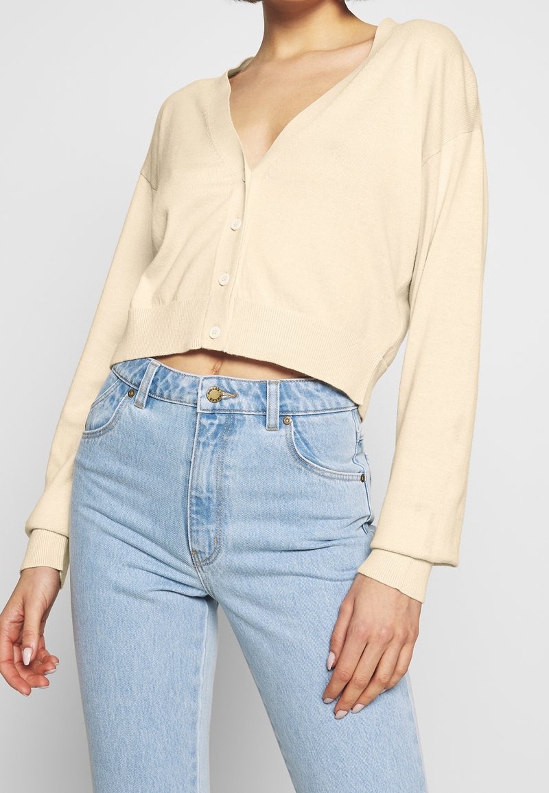 Avkortad beige cardigan med V-ringning, fem knappar och ribbad mudd, som kombineras med högmidjade ljusblå jeans. Mjuk textil, avslappnad passform.
