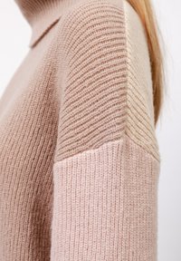 Lichtroze gebreide trui met een hoge kraag, voorzien van een gestructureerd patroon. De mouwen hebben kleurbloksecties in zacht beige.